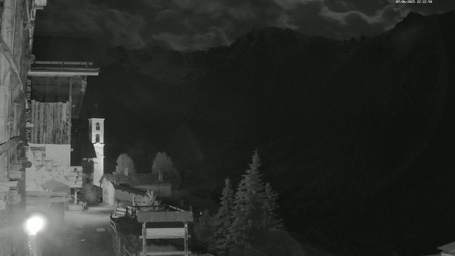 A nighttime, cloudy view of a village nestled in a valley, with a church steeple visible and mountains looming in the background.