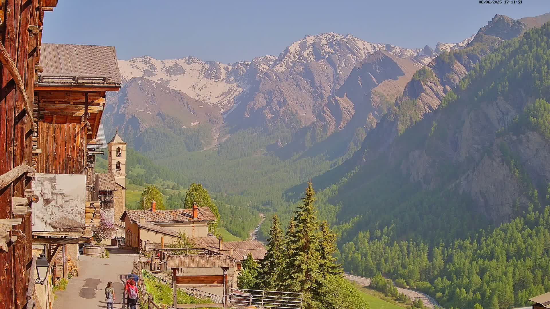 A mountain village with wooden buildings and a church is nestled in a valley, overlooked by snow-capped mountains under a clear, sunny sky.