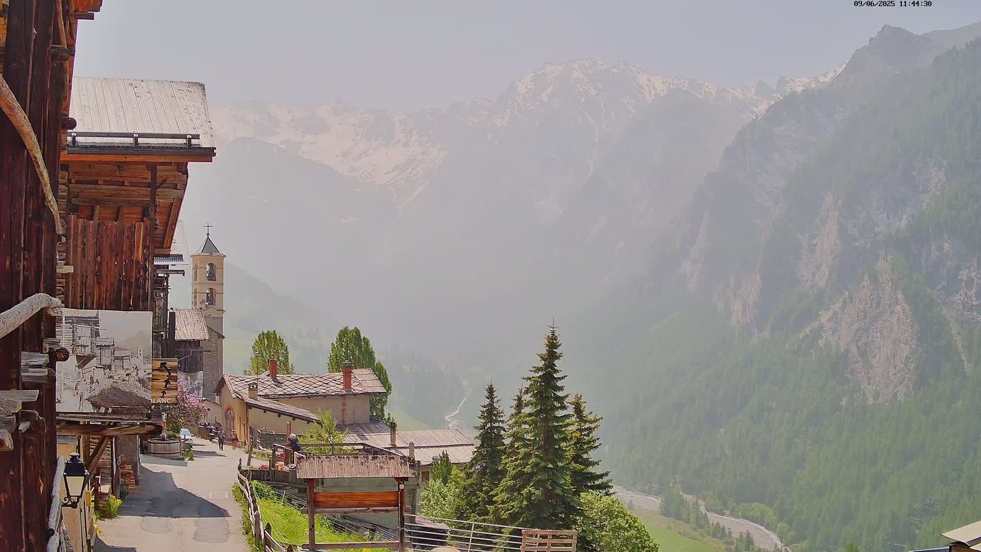 A hazy mountain village with a church steeple, nestled in a valley beneath snow-capped mountains.