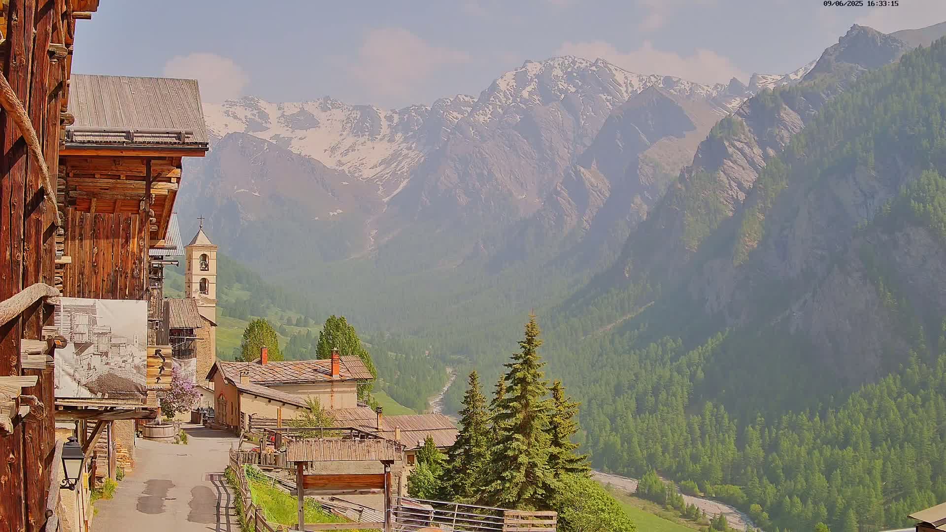 A quaint mountain village with a church nestled in a valley, overlooked by snow-capped mountains under a hazy sunny sky.