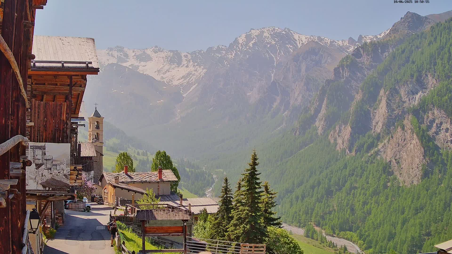 A sunny day reveals a quaint mountain village nestled in a valley, overlooked by snow-capped peaks and lush green forests.