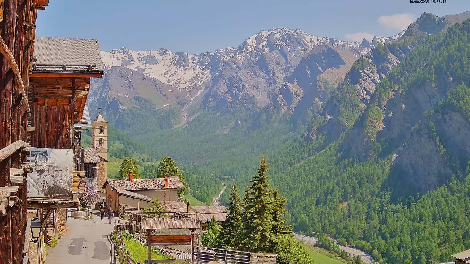 A mountain village with wooden buildings and a church sits nestled in a valley surrounded by snow-capped mountains under a clear, sunny sky.