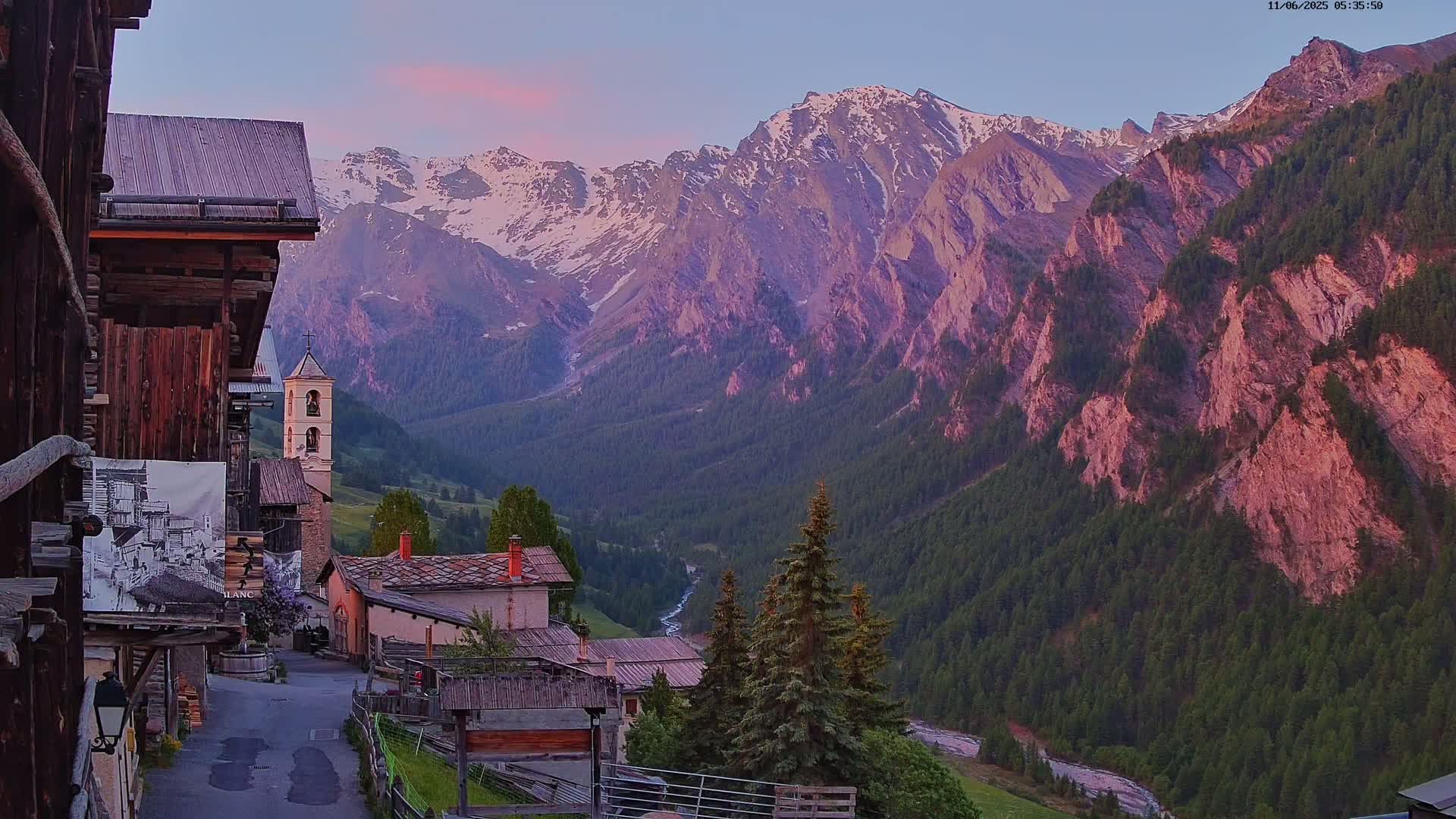 A mountain village with a church steeple is nestled in a valley at sunset, with snow-capped mountains rising in the background under a mostly clear, light-pink sky.