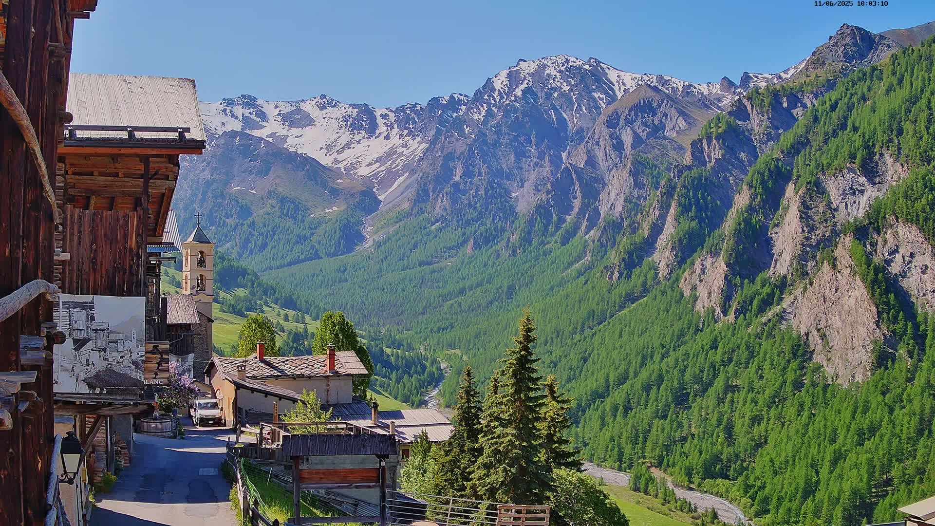 A mountain village nestled in a valley, with a snow-capped mountain range in the background under a clear, sunny sky.