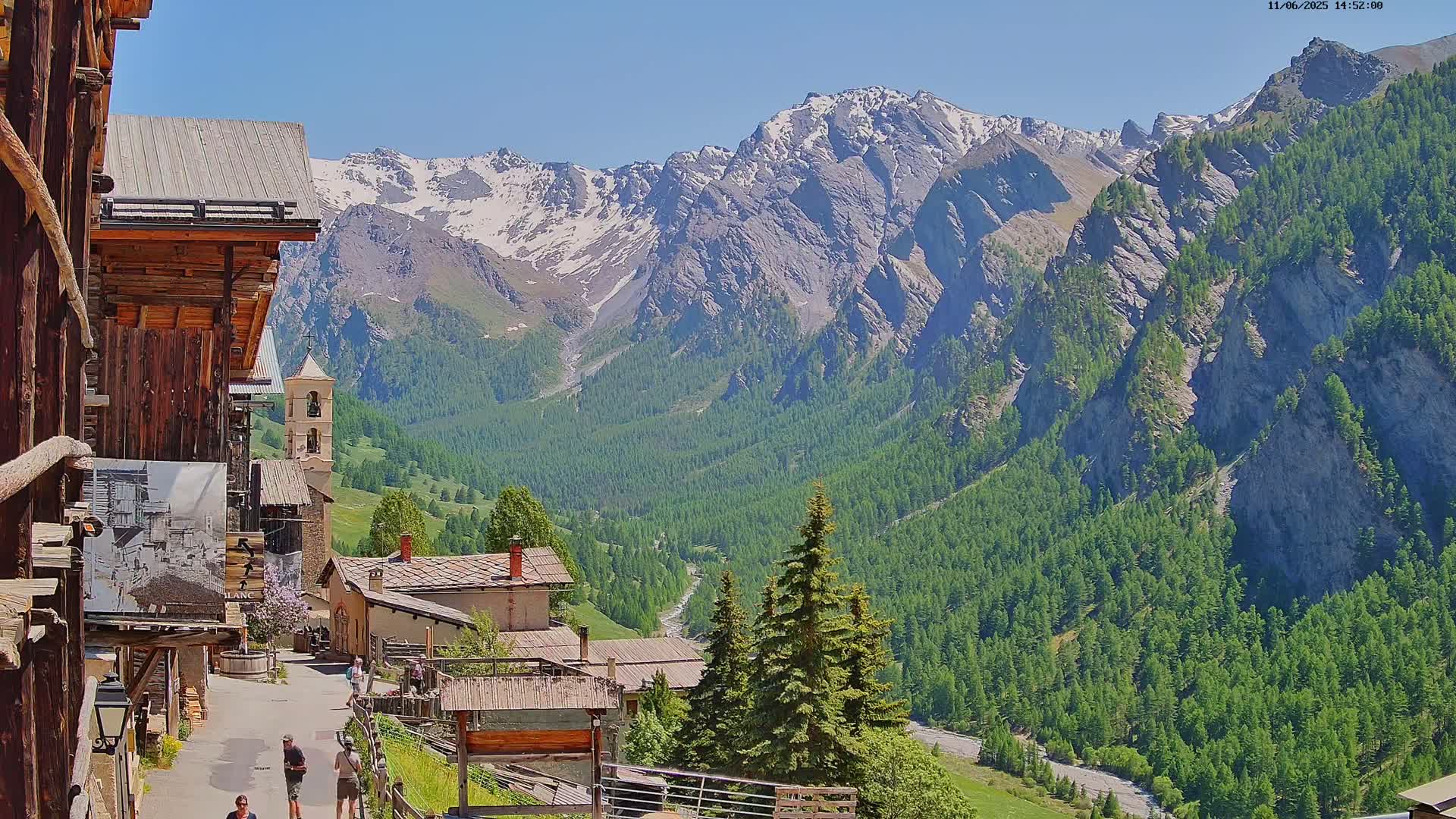A quaint mountain village nestled in a valley, overlooked by snow-capped mountains under a clear, sunny sky.
