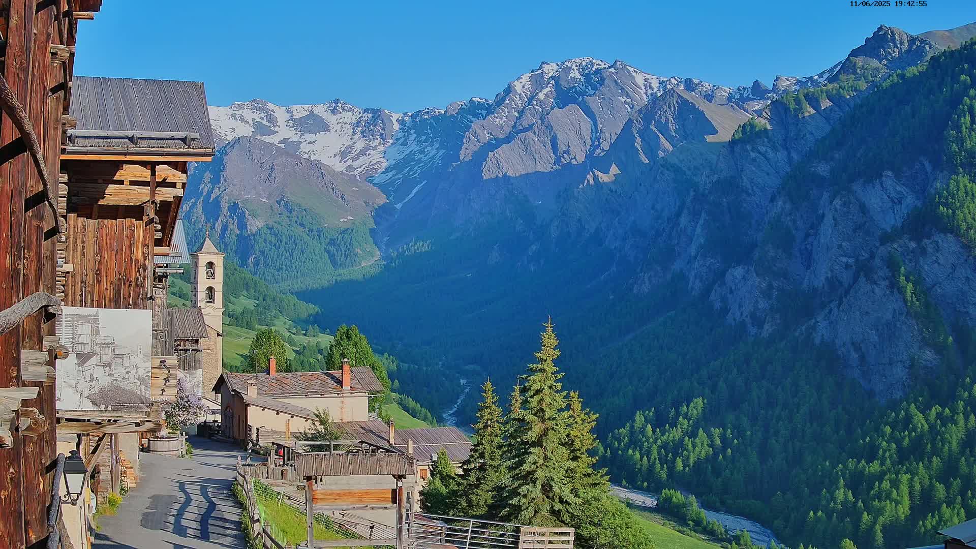 A mountain village with a church nestled in a valley, overlooked by snow-capped mountains under a clear blue sky.