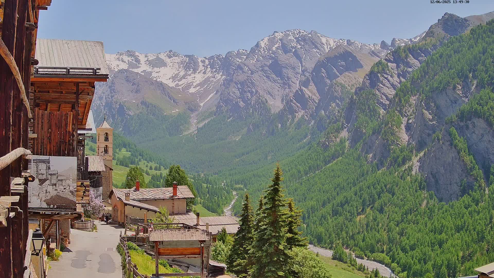 A mountain village with aged wooden buildings nestles in a valley on a sunny day, overlooked by snow-capped peaks and verdant slopes.