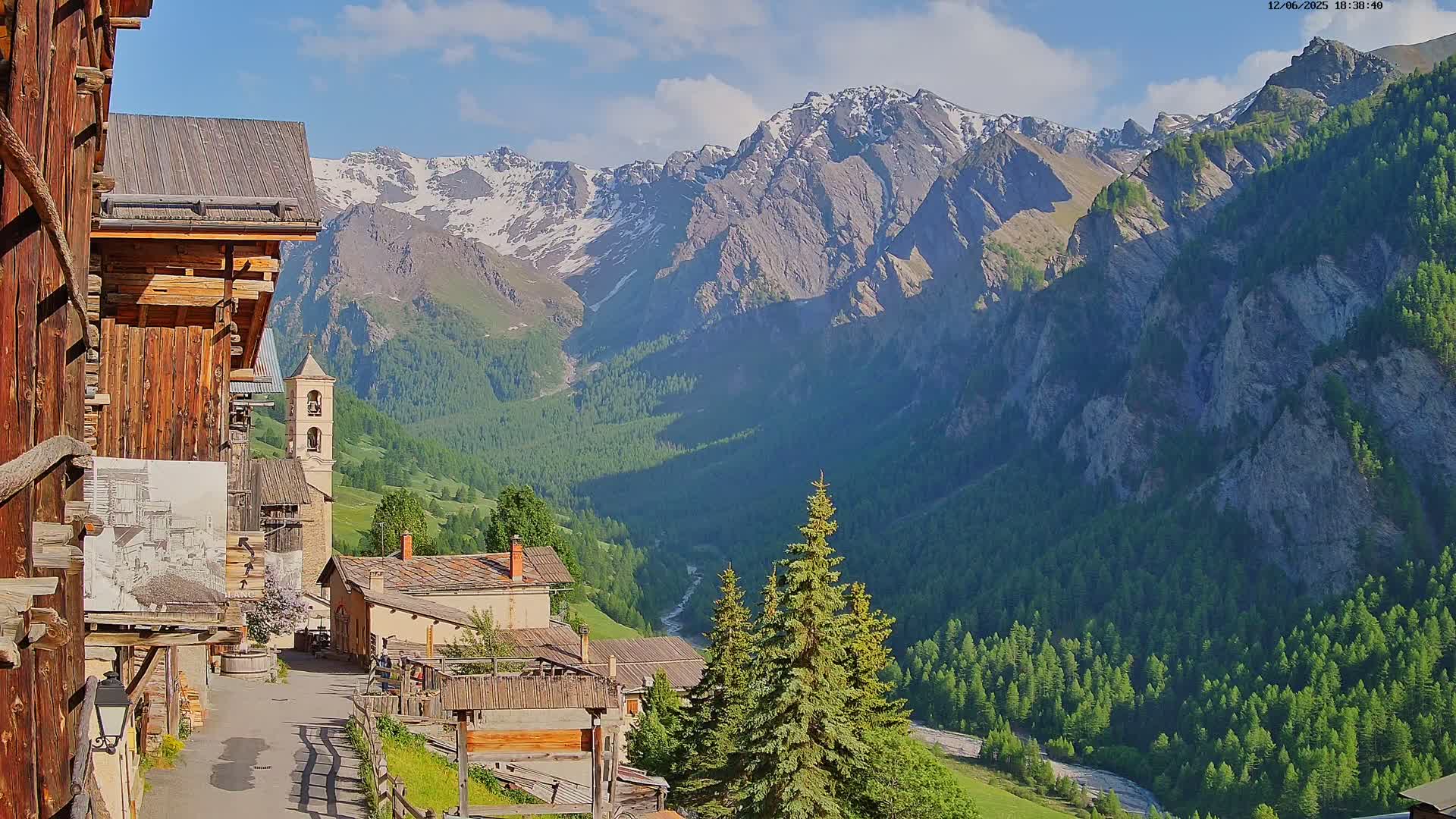A quaint mountain village nestled in a valley, with a snow-capped mountain range in the background under a mostly sunny sky.