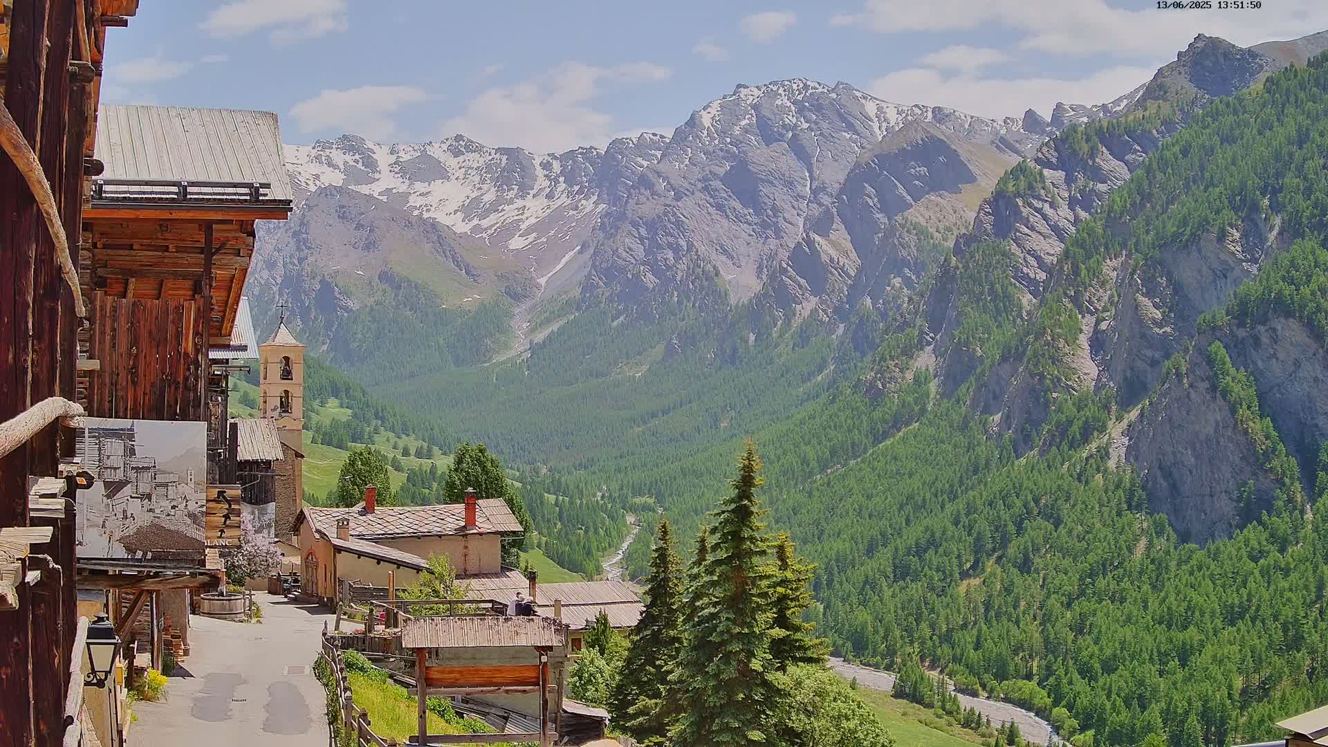 A quaint mountain village with old wooden buildings nestles in a valley, overlooked by snow-capped mountains under a partly sunny sky.