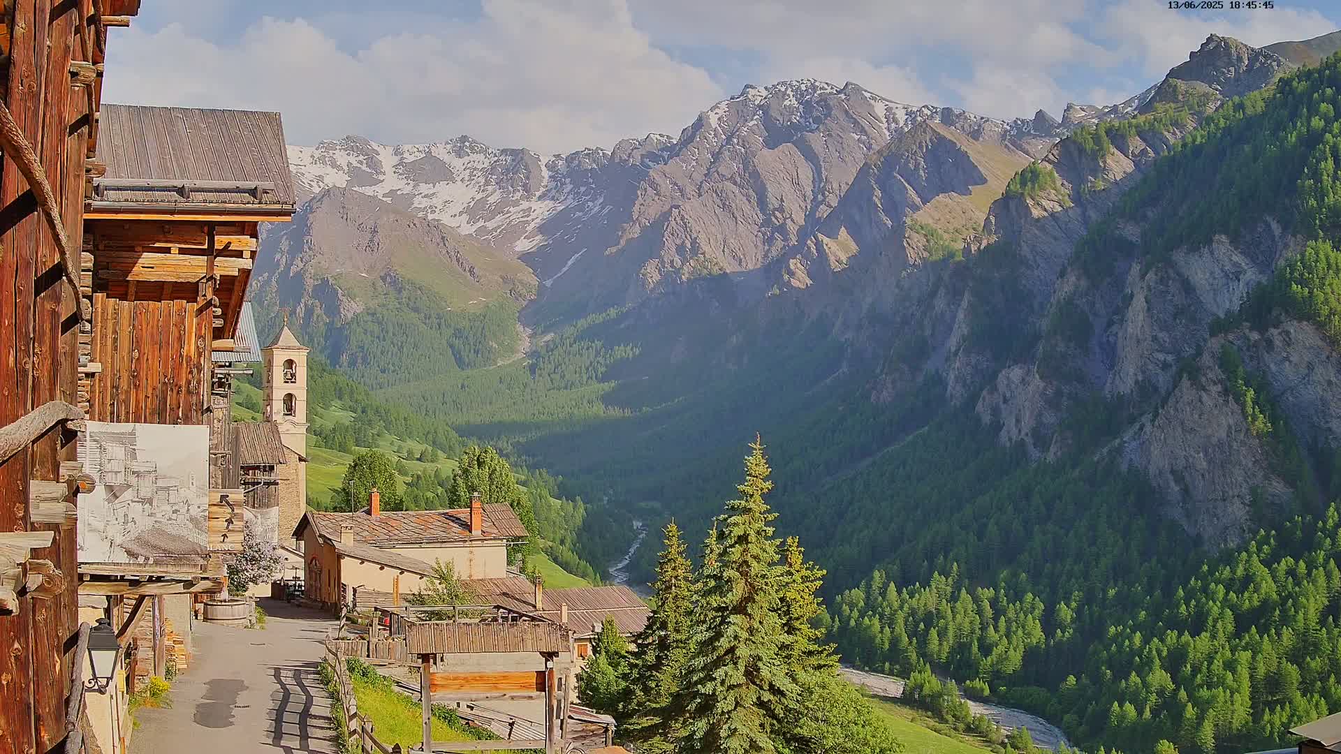A quaint mountain village with wooden buildings nestles in a valley surrounded by snow-capped mountains under a partly cloudy sky.
