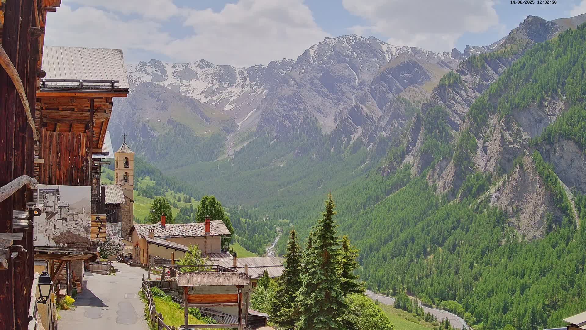 A quaint mountain village with a stone church nestled in a valley, overlooked by snow-capped mountains under a partly cloudy sky.