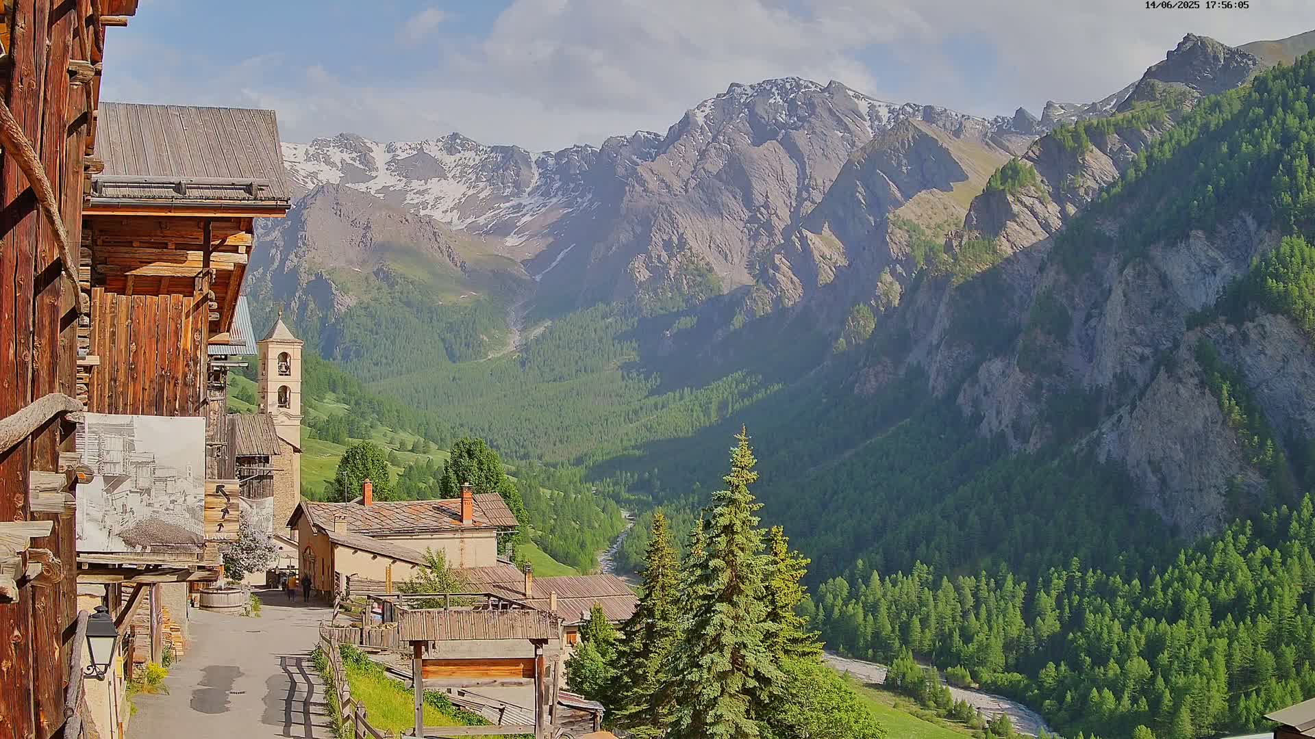 A quaint mountain village with wooden buildings nestles in a valley, overlooked by snow-capped mountains under a mostly sunny sky.