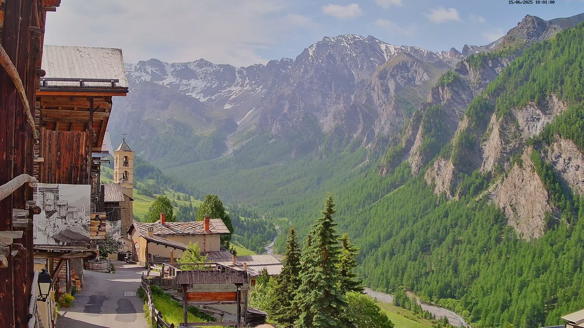 A quaint mountain village with a church steeple nestles in a valley, overlooked by snow-capped mountains under a partly sunny sky.