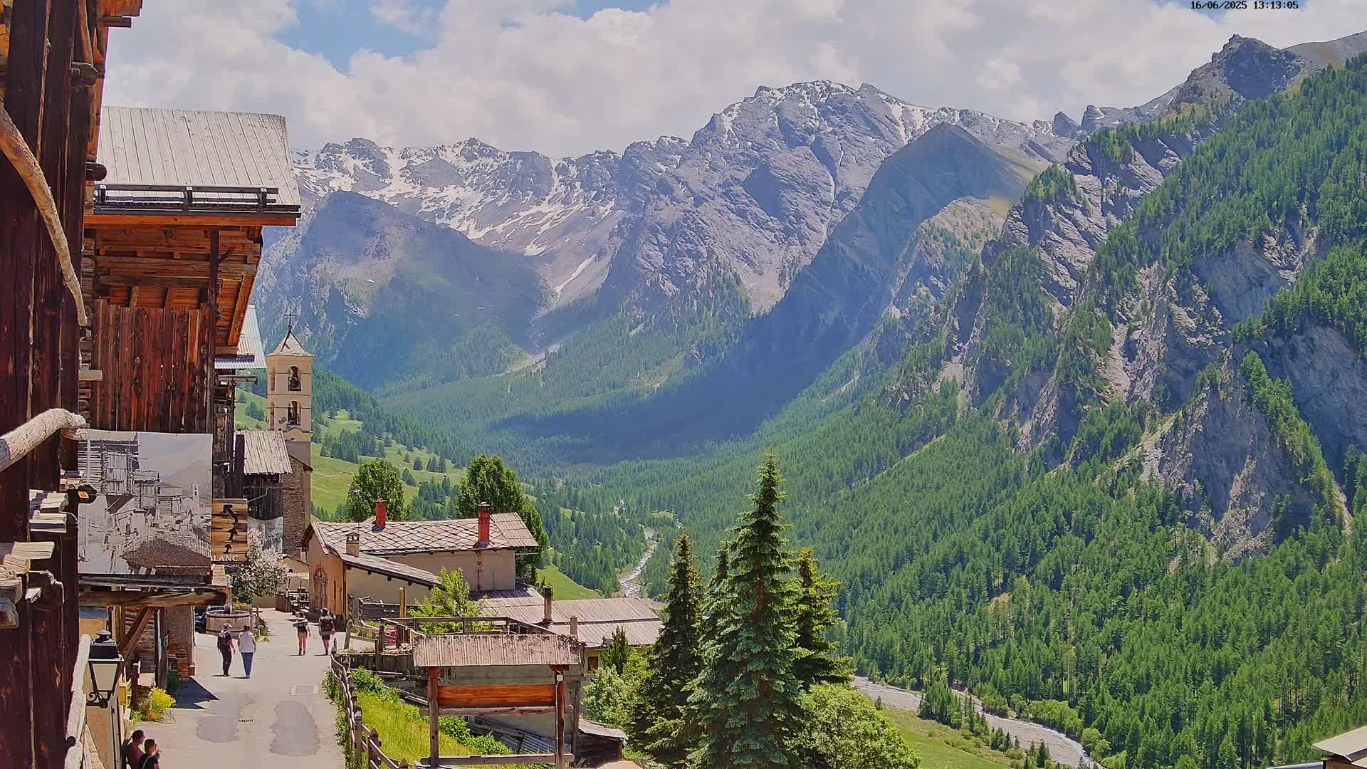 A sunny day reveals a quaint mountain village nestled in a valley, overlooked by snow-capped peaks and lush green forests.