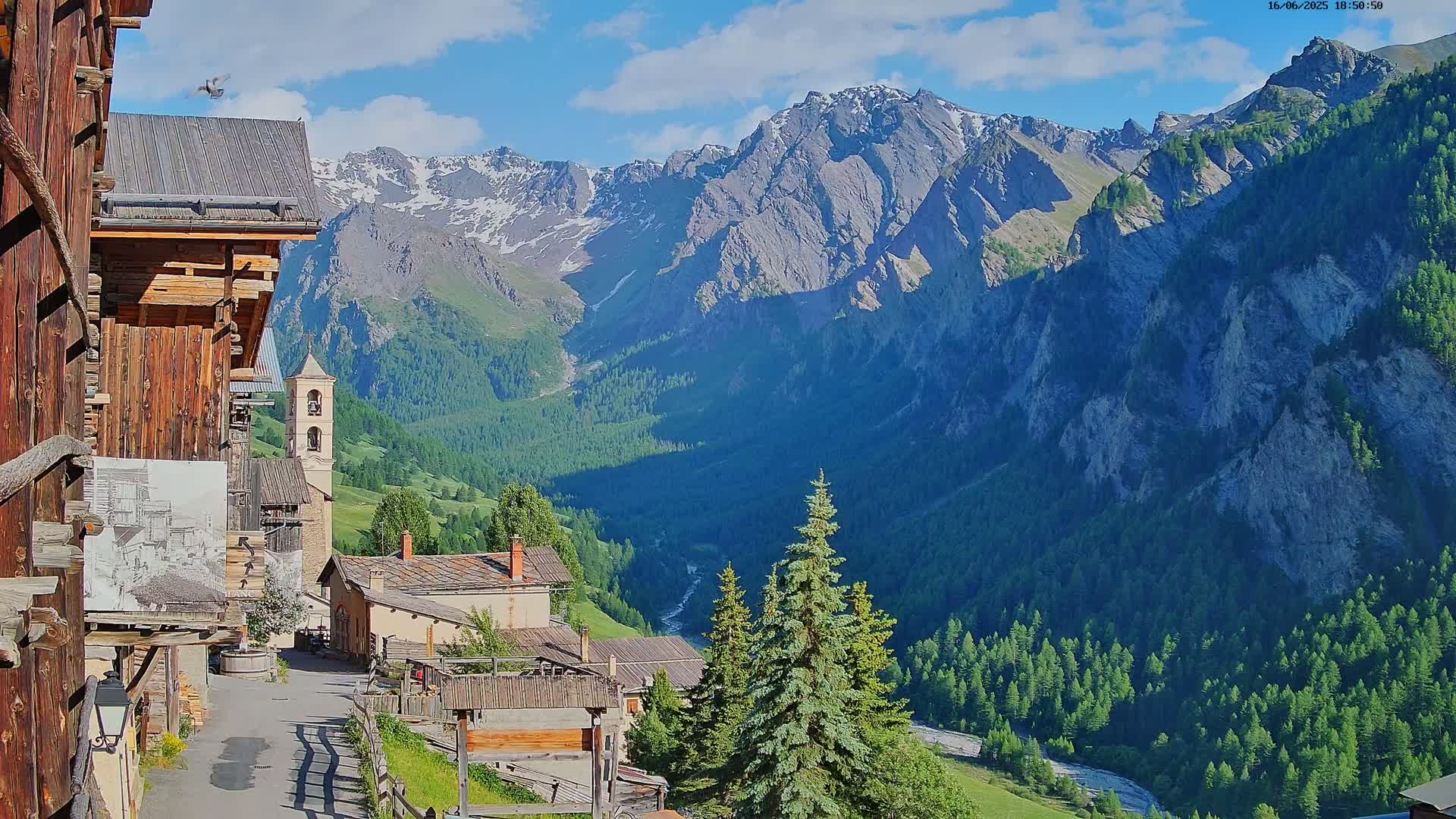 A mountain village with wooden buildings nestles in a valley below snow-capped mountains on a sunny day.