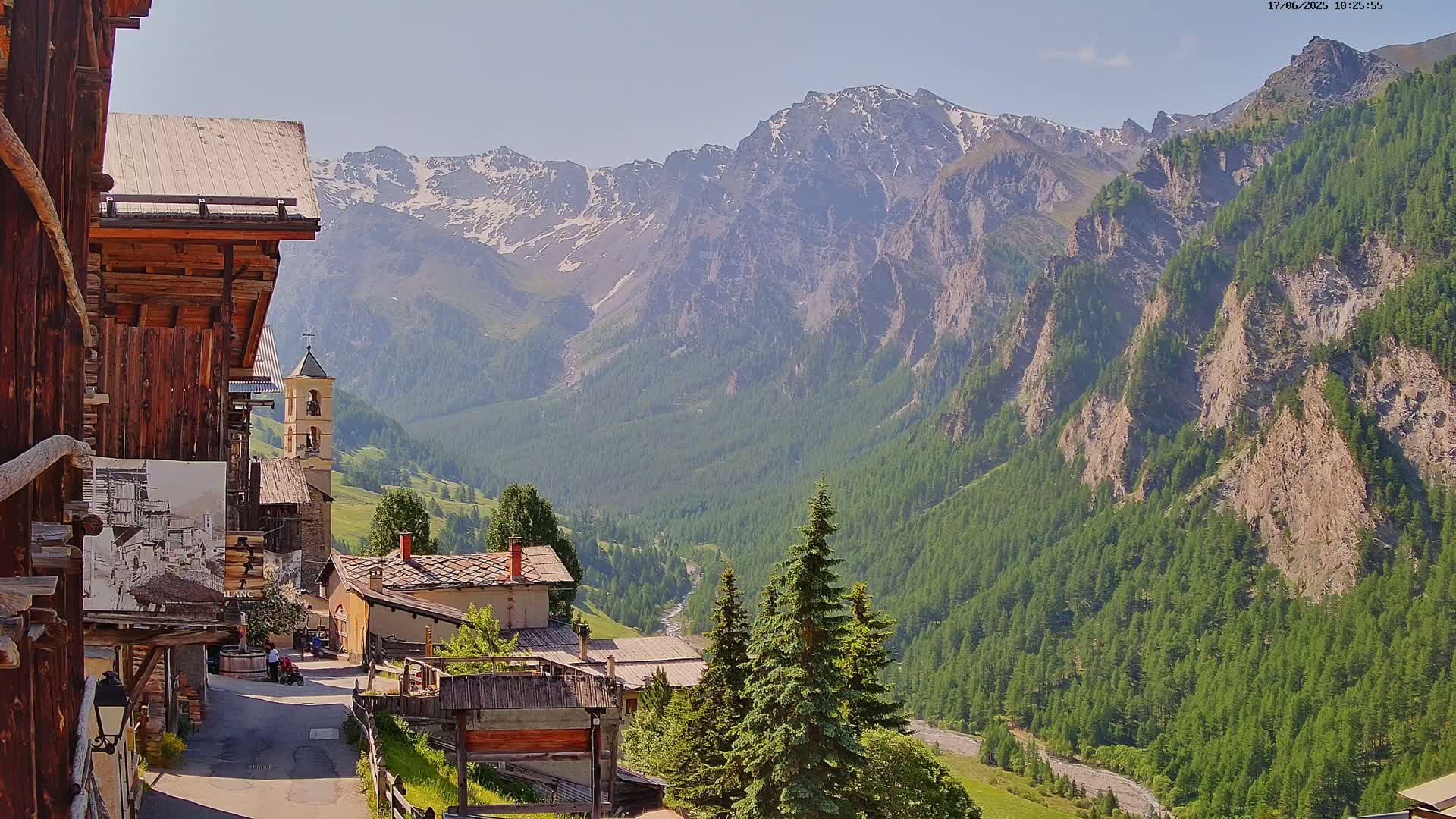 A sunny view of a mountain village nestled in a valley surrounded by snow-capped mountains and lush green forests.
