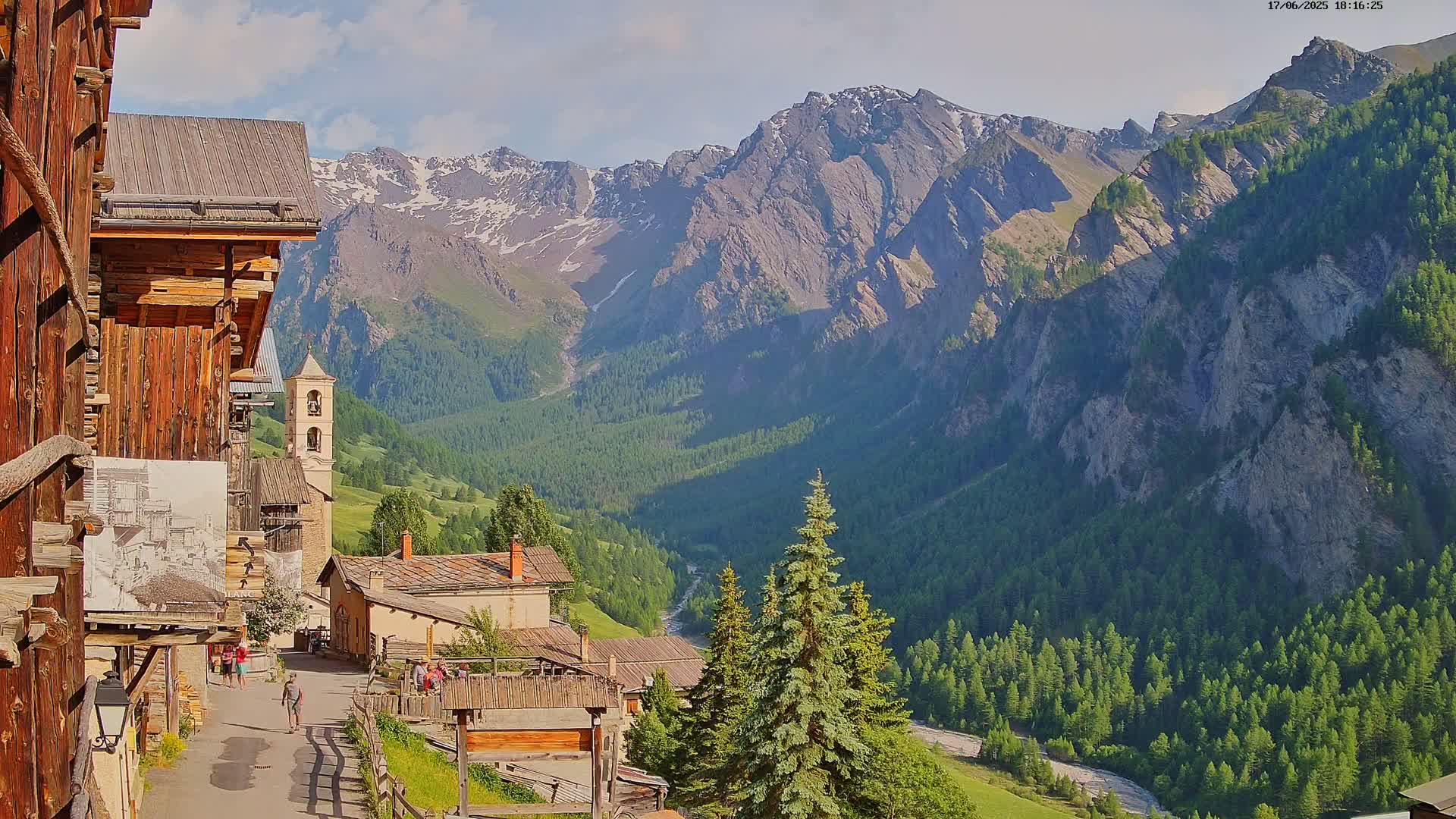 A quaint mountain village with wooden buildings nestles in a valley below snow-capped mountains on a sunny day.