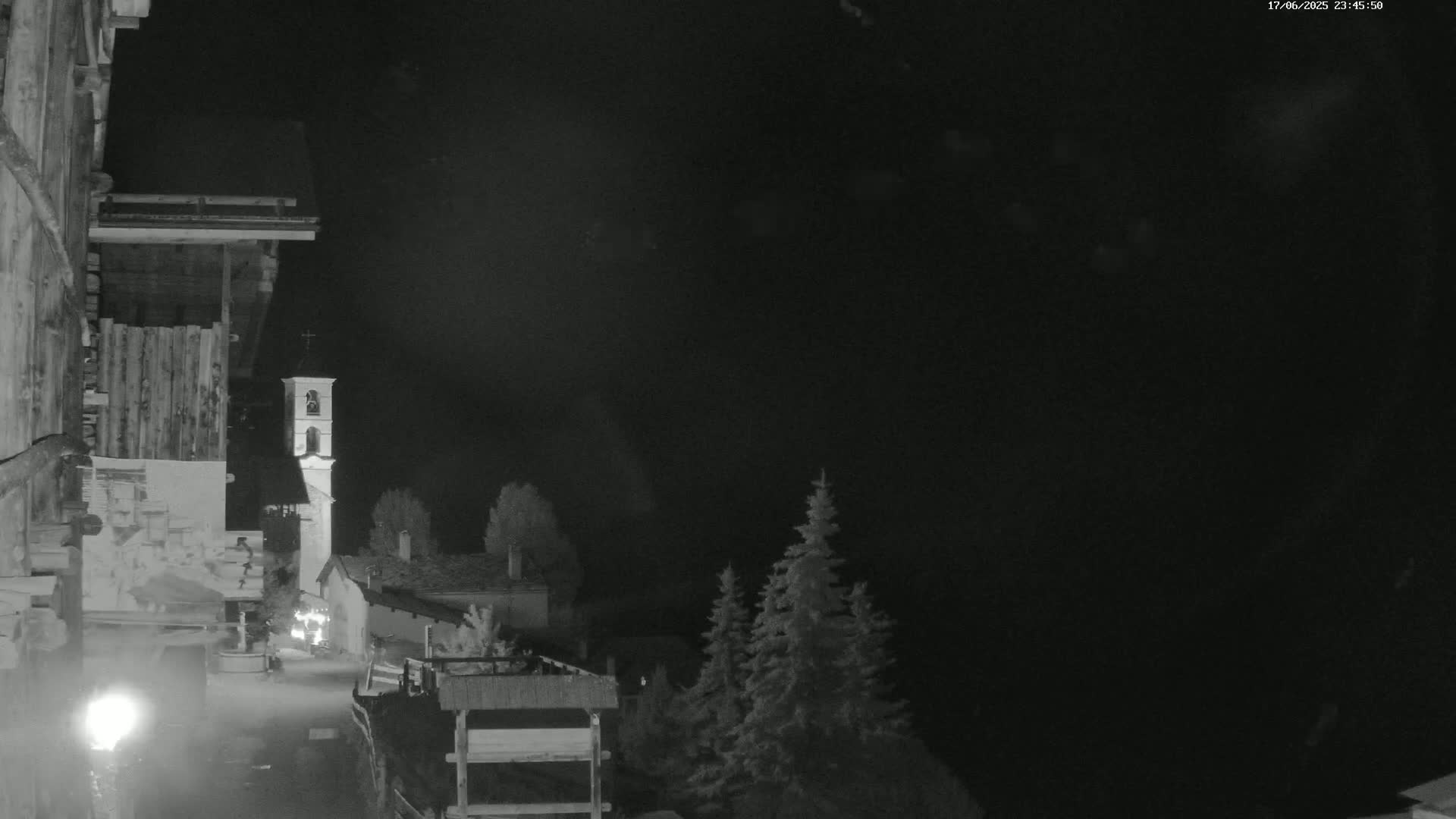A nighttime, black-and-white view of a village with a church steeple, illuminated by a faint light source, under a dark, cloudy sky.
