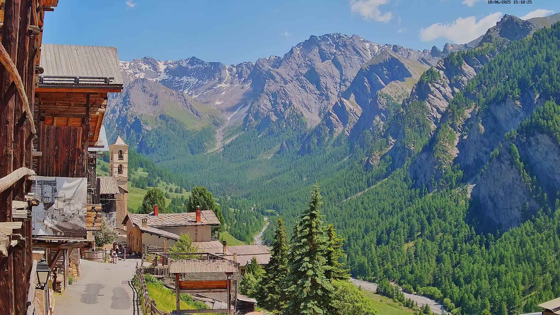 A quaint village nestled in a valley, overlooked by towering, snow-capped mountains under a bright, sunny sky.
