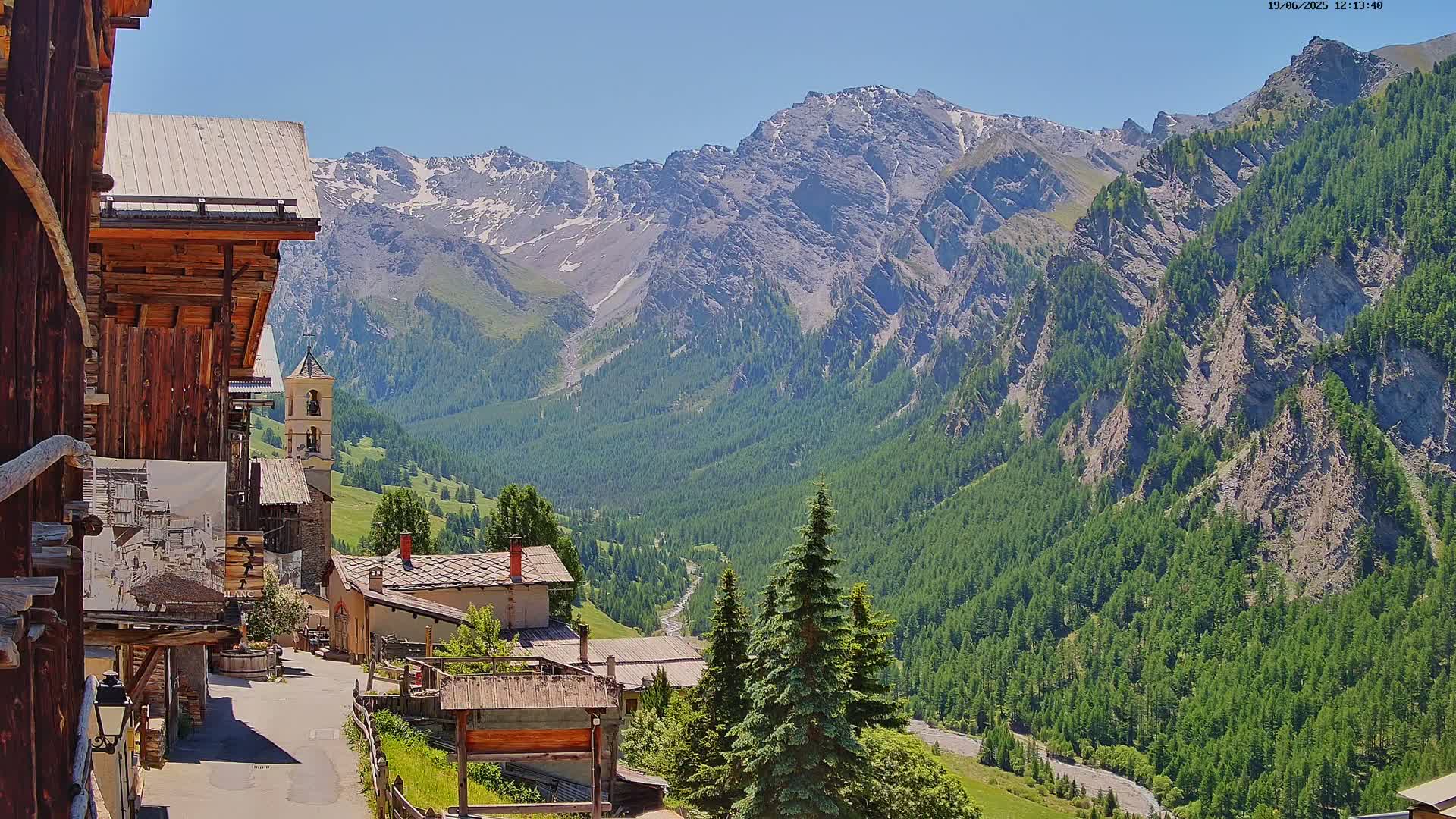 A sunny mountain village nestled in a valley, surrounded by lush green forests and snow-capped peaks.