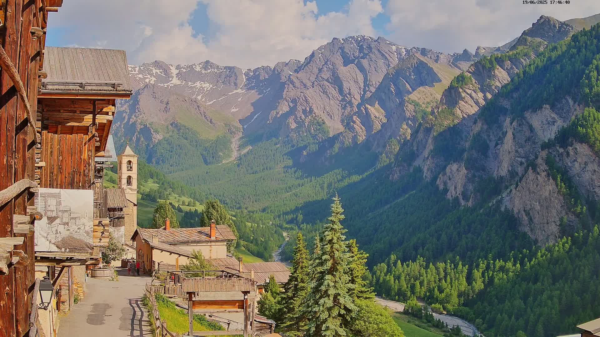A mountain village with wooden buildings nestled in a valley, overlooked by a range of snow-capped mountains under a partly sunny sky.