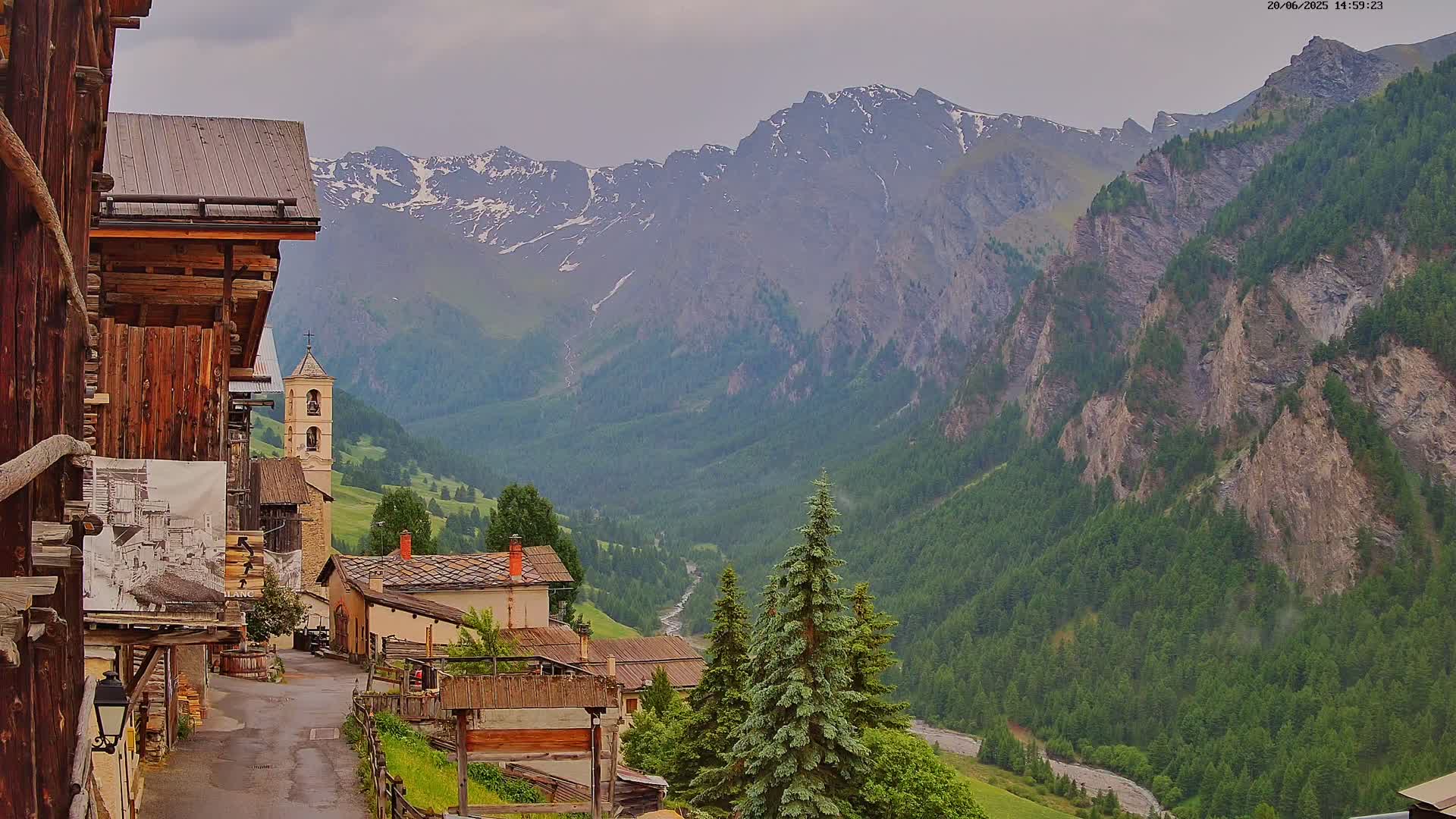 A mountain village nestled in a valley, overlooked by snow-capped peaks under an overcast sky.