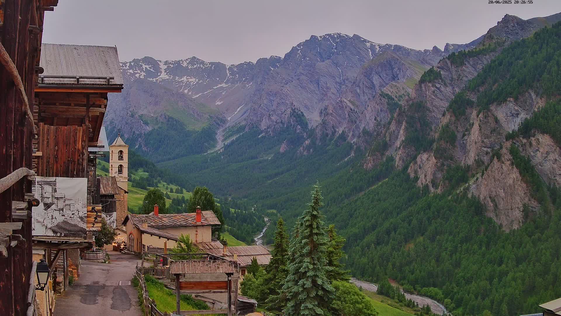 A quaint mountain village with a church steeple is nestled in a valley surrounded by towering, snow-capped mountains under an overcast sky.