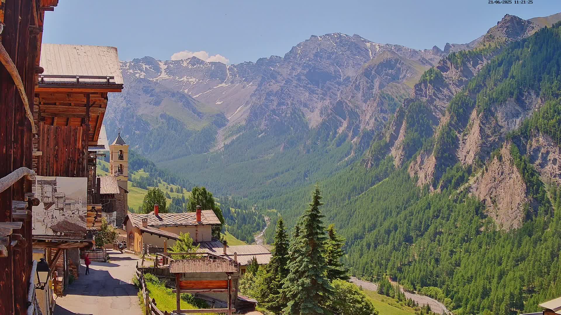 A quaint mountain village nestled in a valley, surrounded by lush green forests and towering snow-capped mountains under a sunny sky.