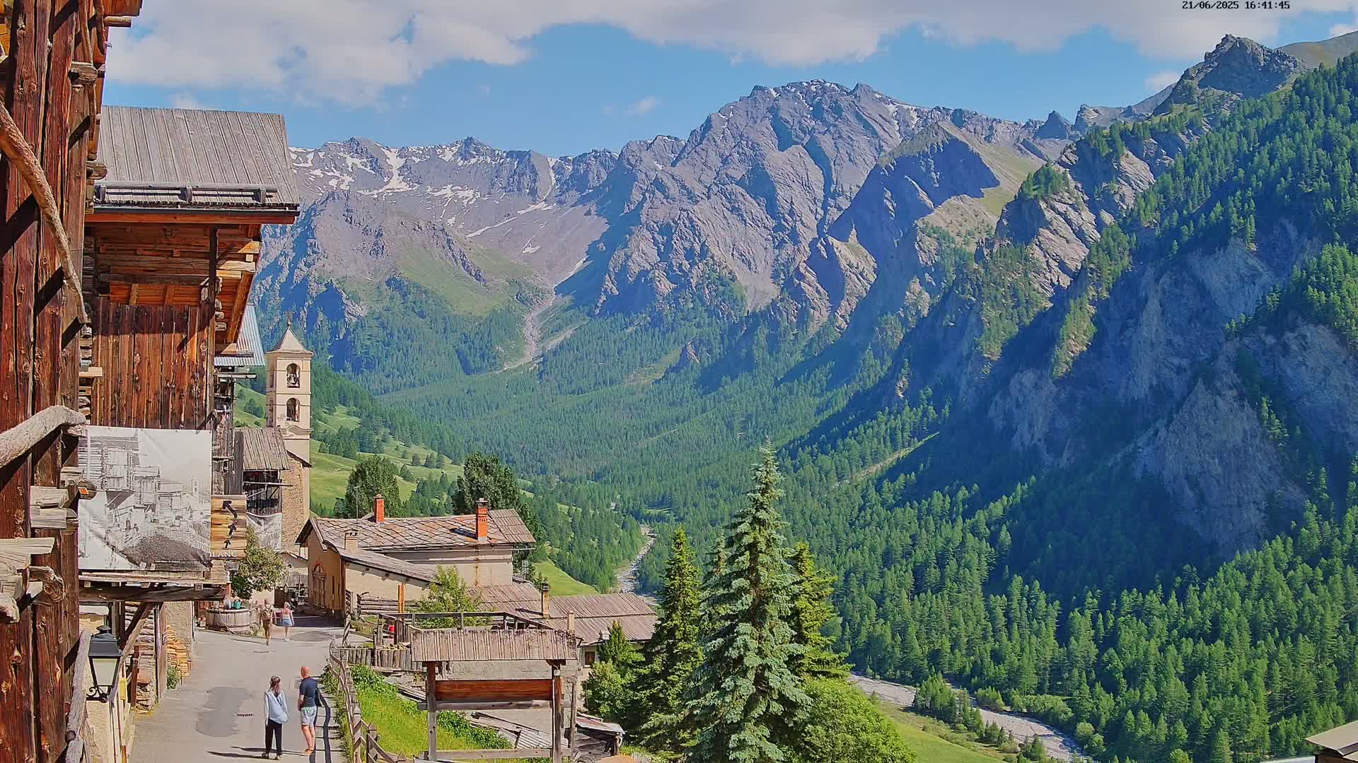 A mountain village nestled in a valley, under a partly sunny sky, is surrounded by steep, rocky mountains covered in evergreen trees.
