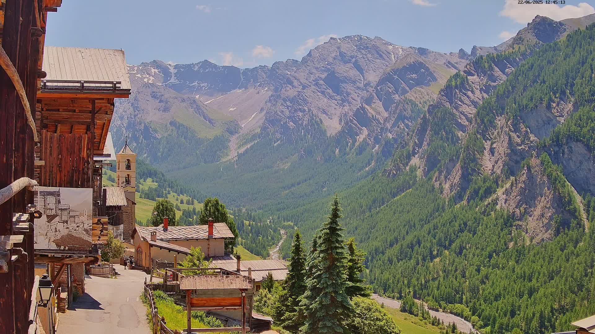 A mountain village nestled in a valley, with a church steeple visible, overlooks a vast, sun-drenched mountainous landscape covered in green pine forests under a mostly clear sky.