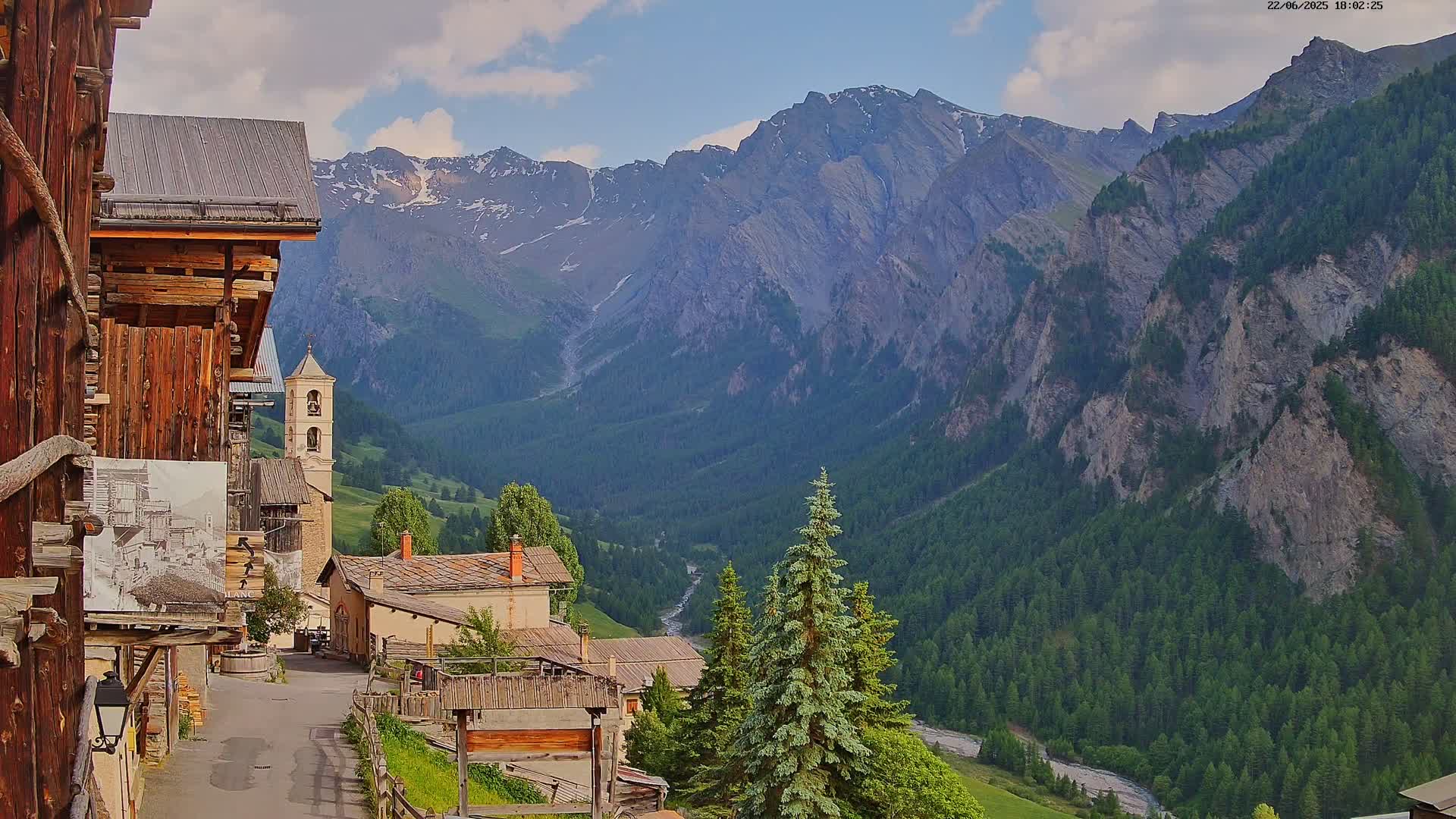 A quaint mountain village with a church steeple nestles in a valley surrounded by towering, rocky mountains under a partly cloudy sky.