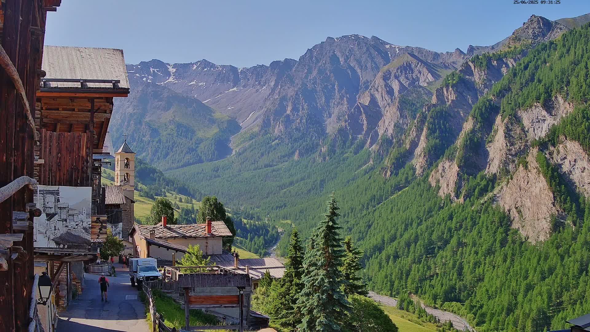 A sunny mountain valley village scene shows a stone church steeple, wooden buildings, and a roadway leading to a mountain range with a river at its base.