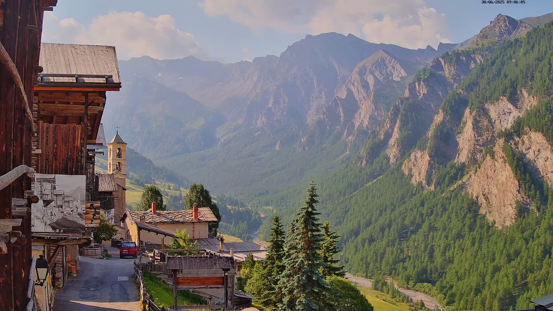 A quaint village nestled in a valley, surrounded by towering mountains under a partly cloudy sky.