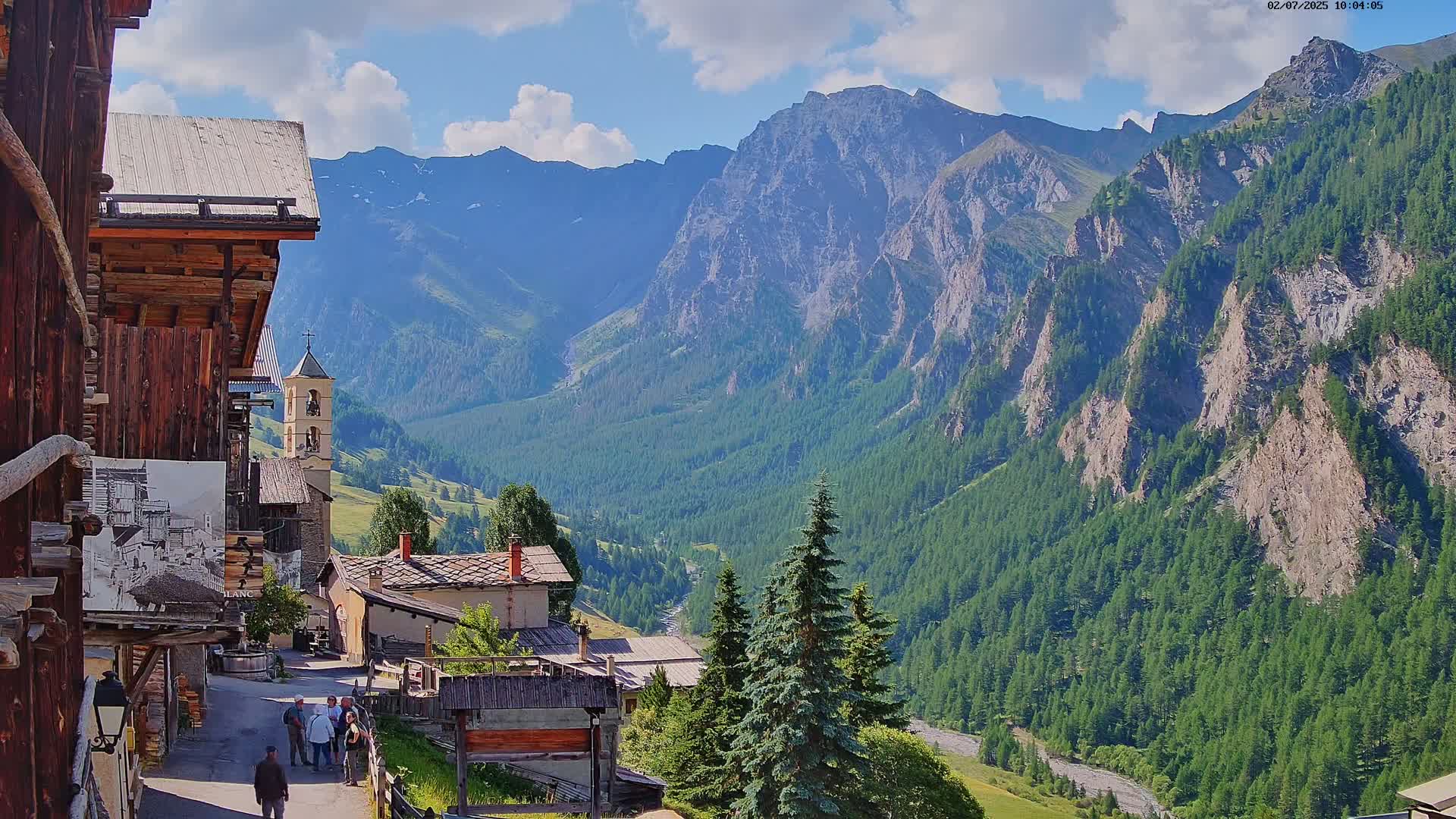 A quaint mountain village nestled in a valley, overlooked by towering, sunlit mountains under a partly cloudy sky.