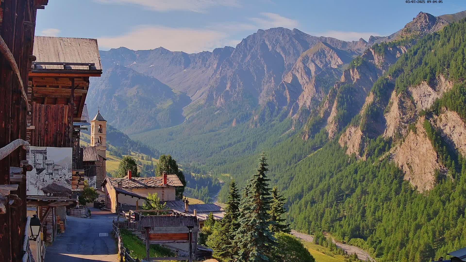 A mountain village with old wooden buildings and a church overlooks a valley of green forests and steep, rocky mountains under a mostly sunny sky.