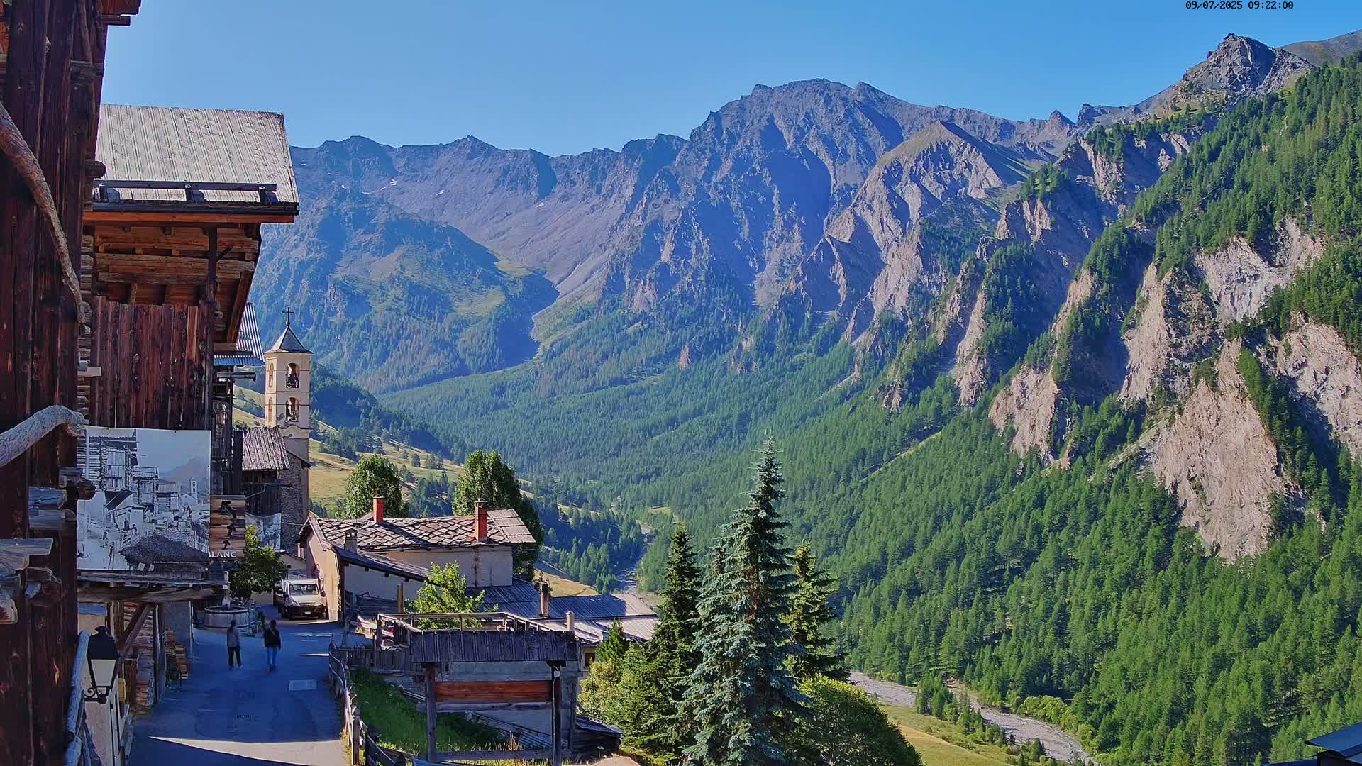 A mountain village with a church nestled in a valley, surrounded by steep, green, rocky mountains under a clear blue sky.