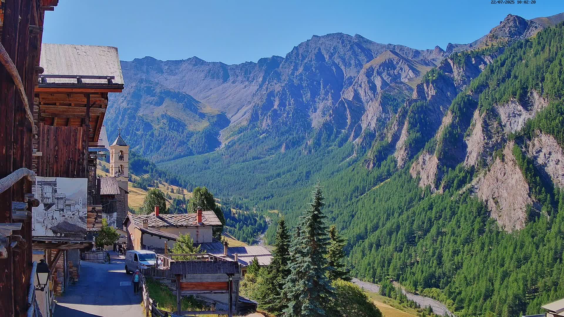 A mountain village nestled in a valley, surrounded by steep, rocky mountains covered in evergreen trees, under a clear blue sky.