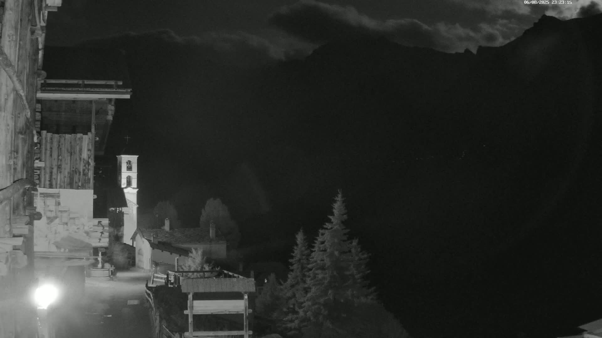 A nighttime view of a small village nestled in a valley, illuminated by a single lamppost, with a church steeple visible and dark, cloudy skies above mountains in the background.