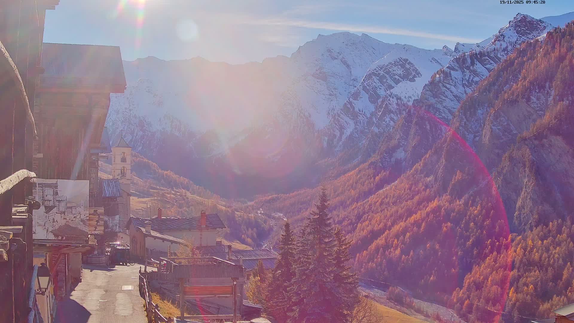 A bright sunny day illuminates a charming mountain village with a visible church bell tower, nestled in a valley vibrant with golden autumn foliage, all framed by majestic snow-capped peaks under a clear sky.