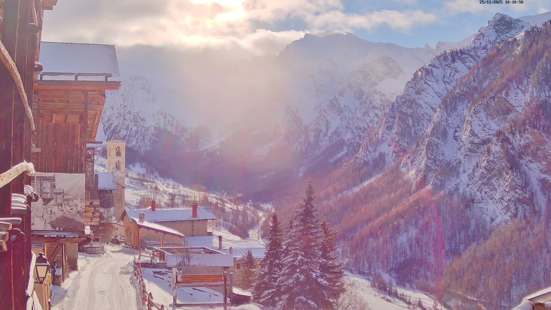 A picturesque snowy mountain village, featuring traditional wooden buildings and a prominent church tower along a winding path, is set against a backdrop of towering, snow-covered mountains and a valley of bare trees, under a partially cloudy sky with bright sunlight creating a warm, hazy glow.