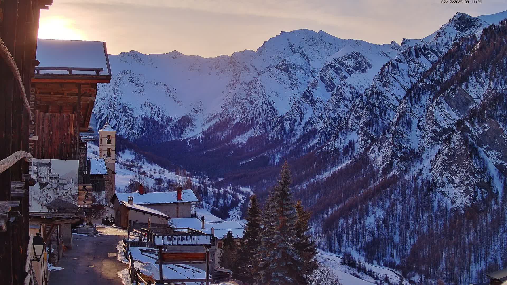 A snow-covered village featuring a church tower and traditional wooden buildings is bathed in early morning light against a backdrop of vast, rugged, snow-laden mountains and dark forested valleys under a clear, cold sky.