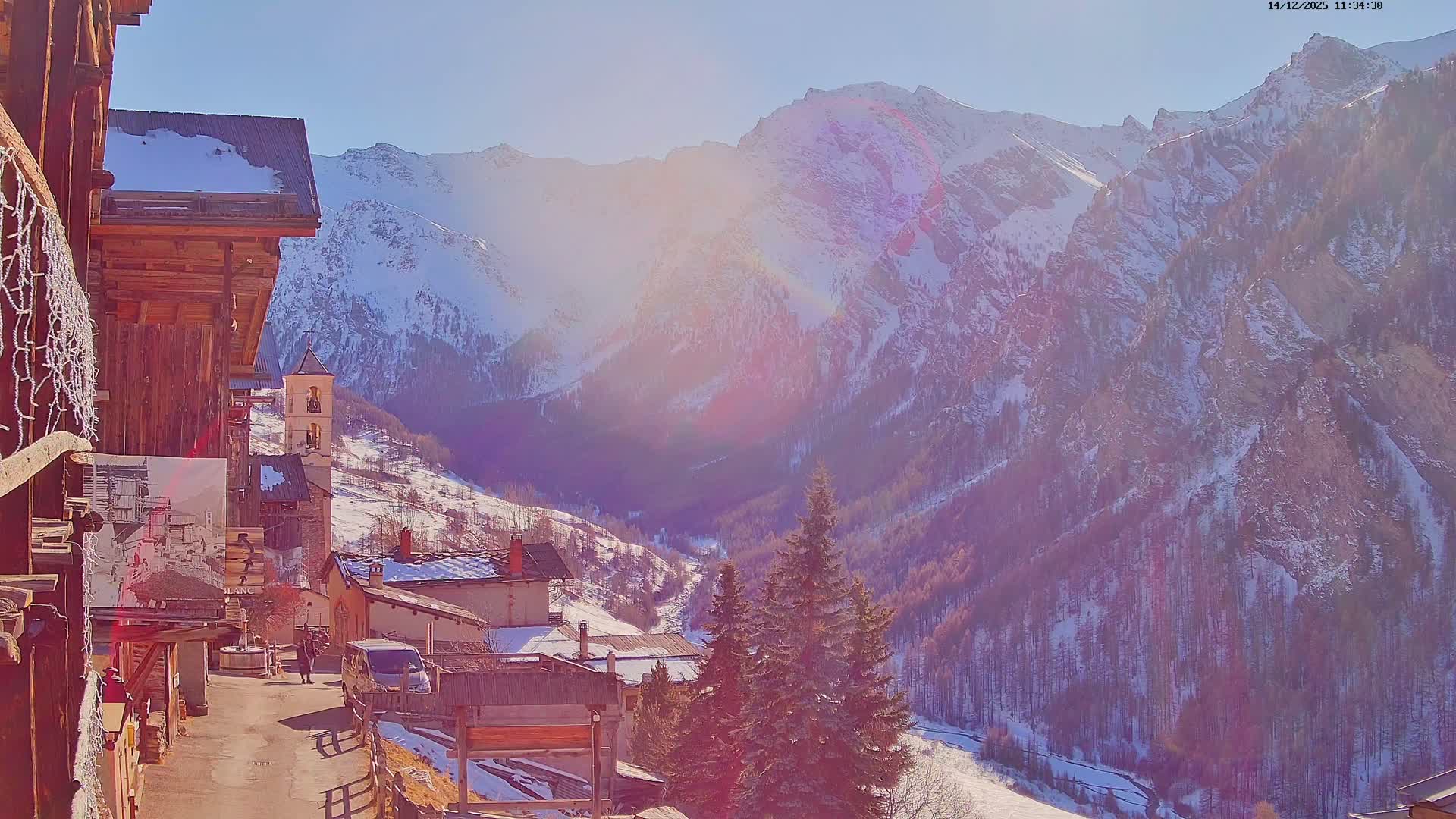 A snow-covered village featuring a church tower and traditional wooden buildings is bathed in early morning light against a backdrop of vast, rugged, snow-laden mountains and dark forested valleys under a clear, cold sky.