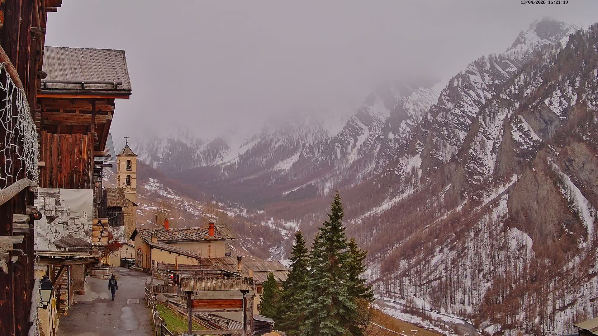 A snow-covered village featuring a church tower and traditional wooden buildings is bathed in early morning light against a backdrop of vast, rugged, snow-laden mountains and dark forested valleys under a clear, cold sky.
