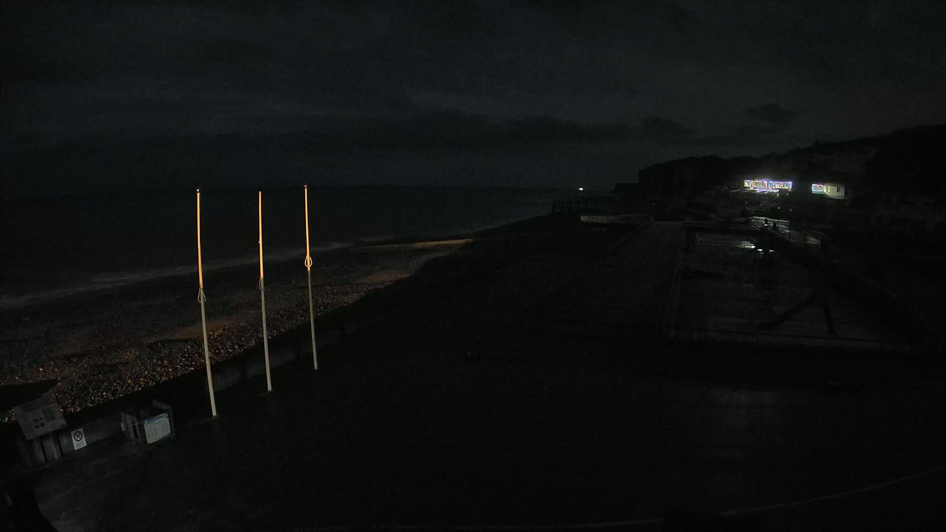 At night, three illuminated poles stand near a dark, pebble-covered beach under a cloudy sky, with faintly lit buildings visible on a cliff in the distance.