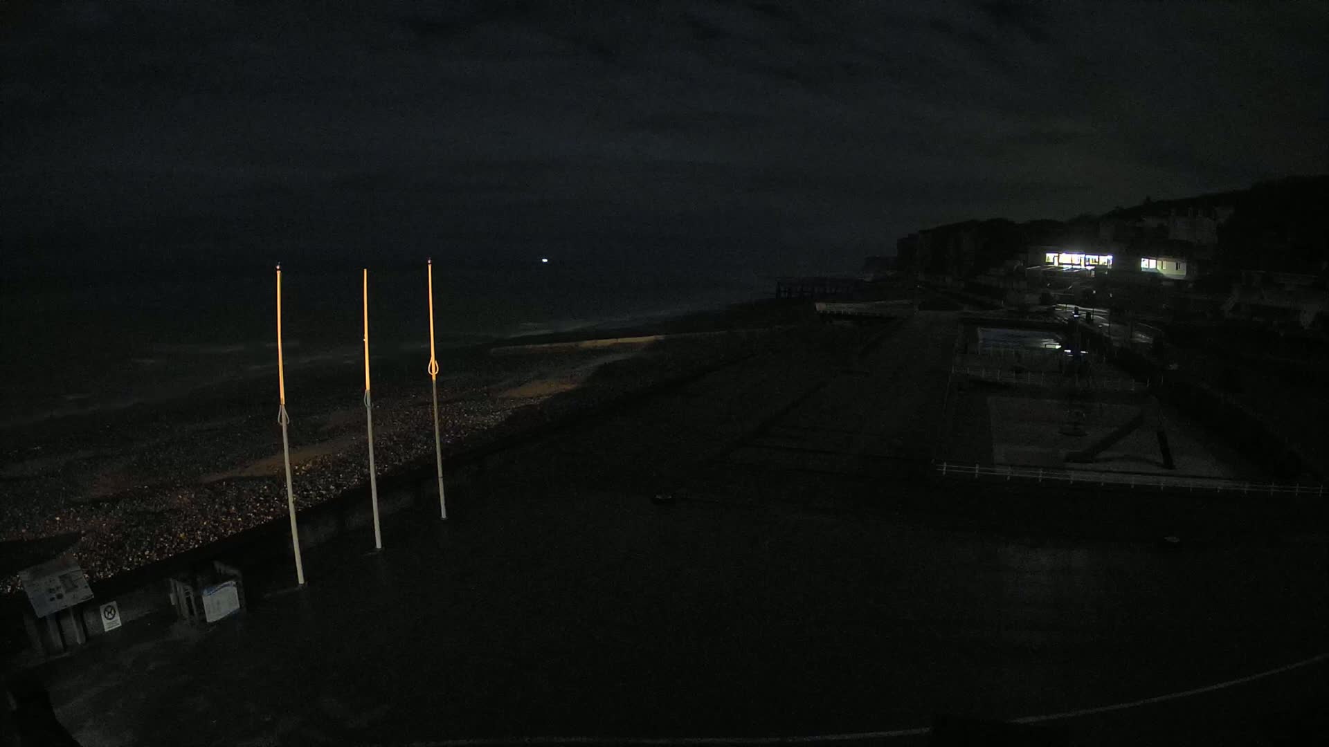 At night, three tall, slender lights stand on a dark, pebble-strewn beach near a dimly lit building and a pier under a mostly clear sky.