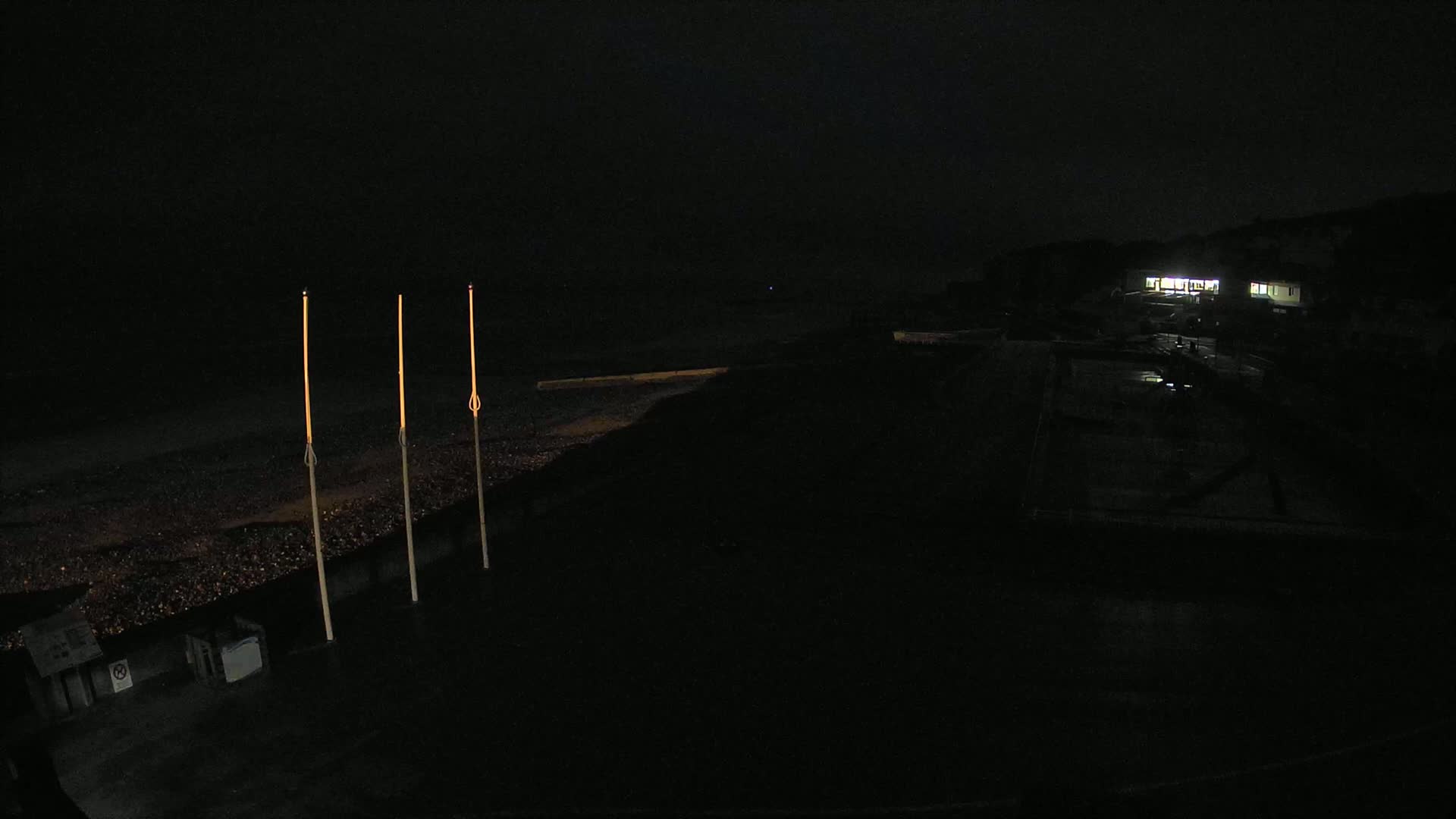 Three tall, slender lights illuminate a dark, pebble-strewn beach at night, with a dimly lit building visible in the distance.