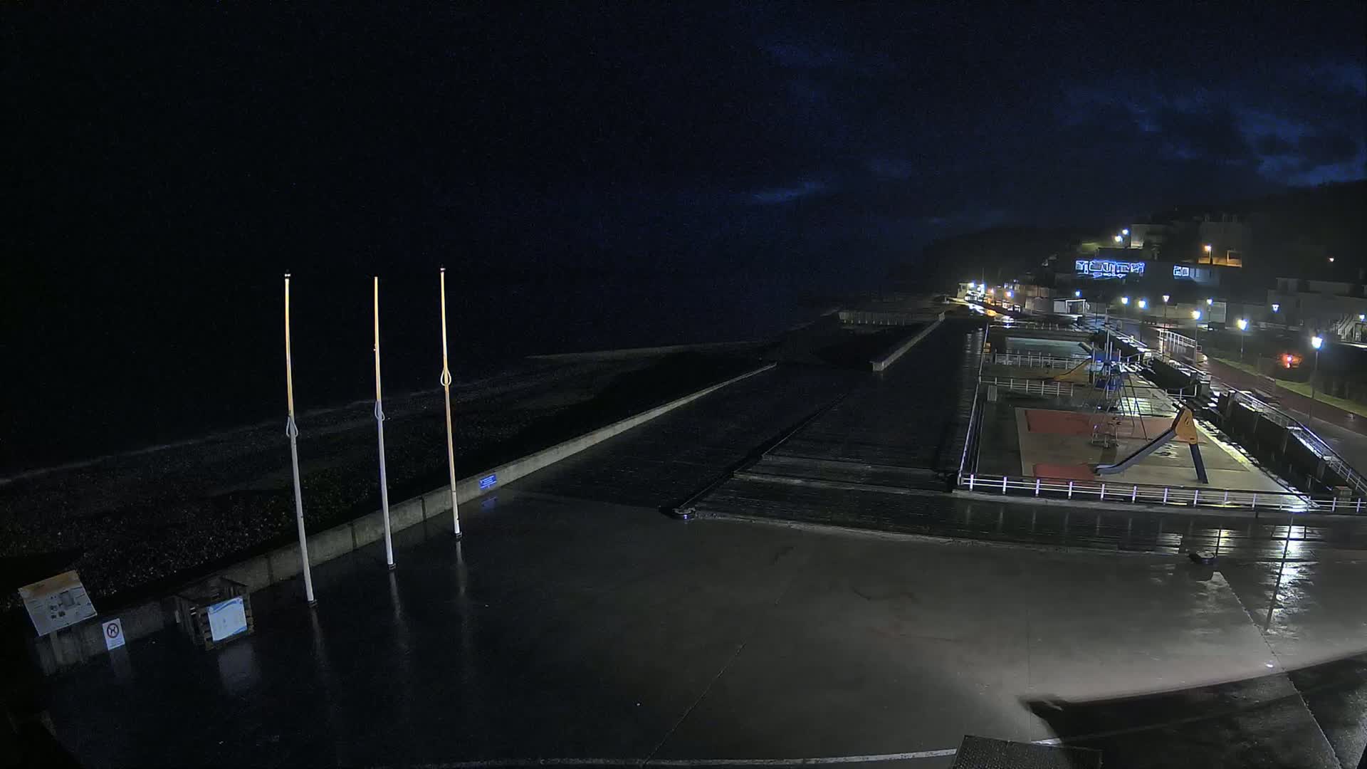 A rain-slicked, nighttime seaside promenade features a playground, a dark ocean, and buildings on a hill in the distance.