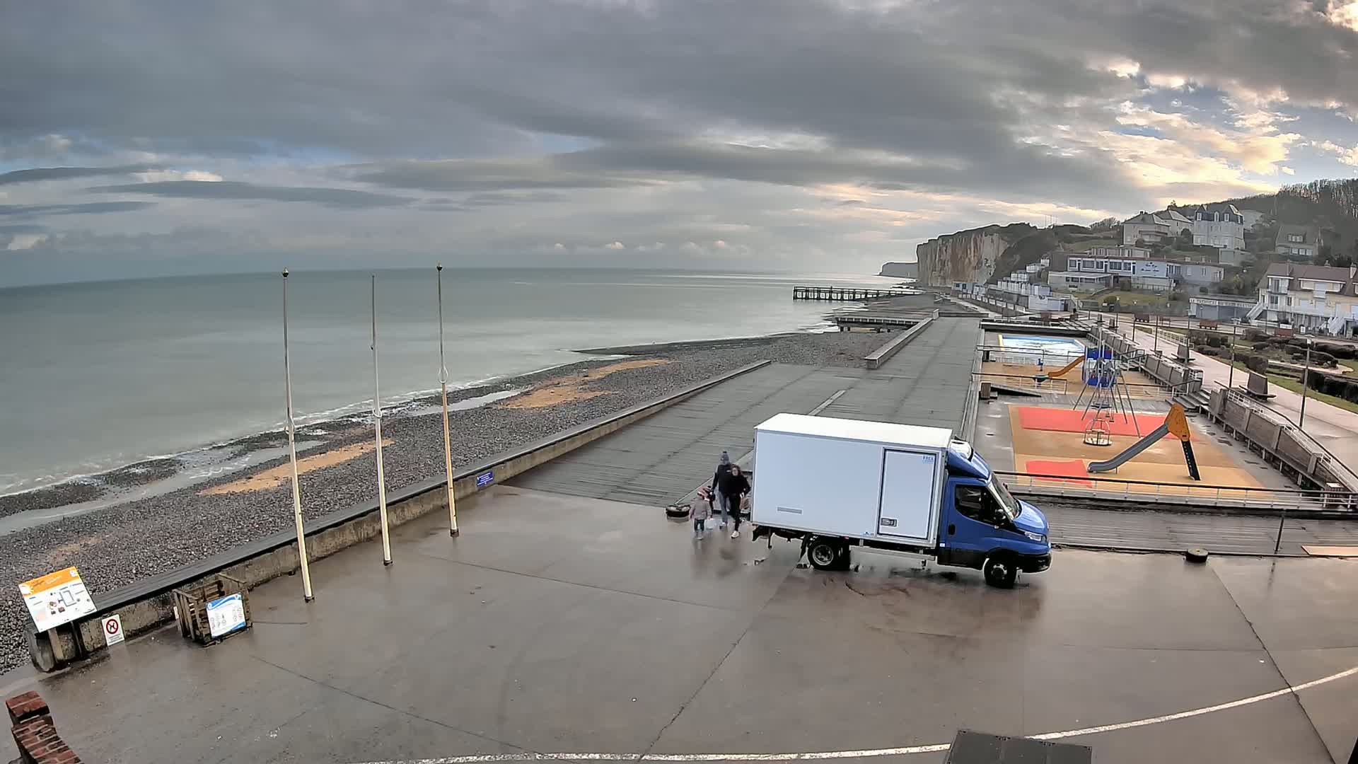 A blue van is parked near a playground by the ocean on an overcast day, with a family walking nearby.