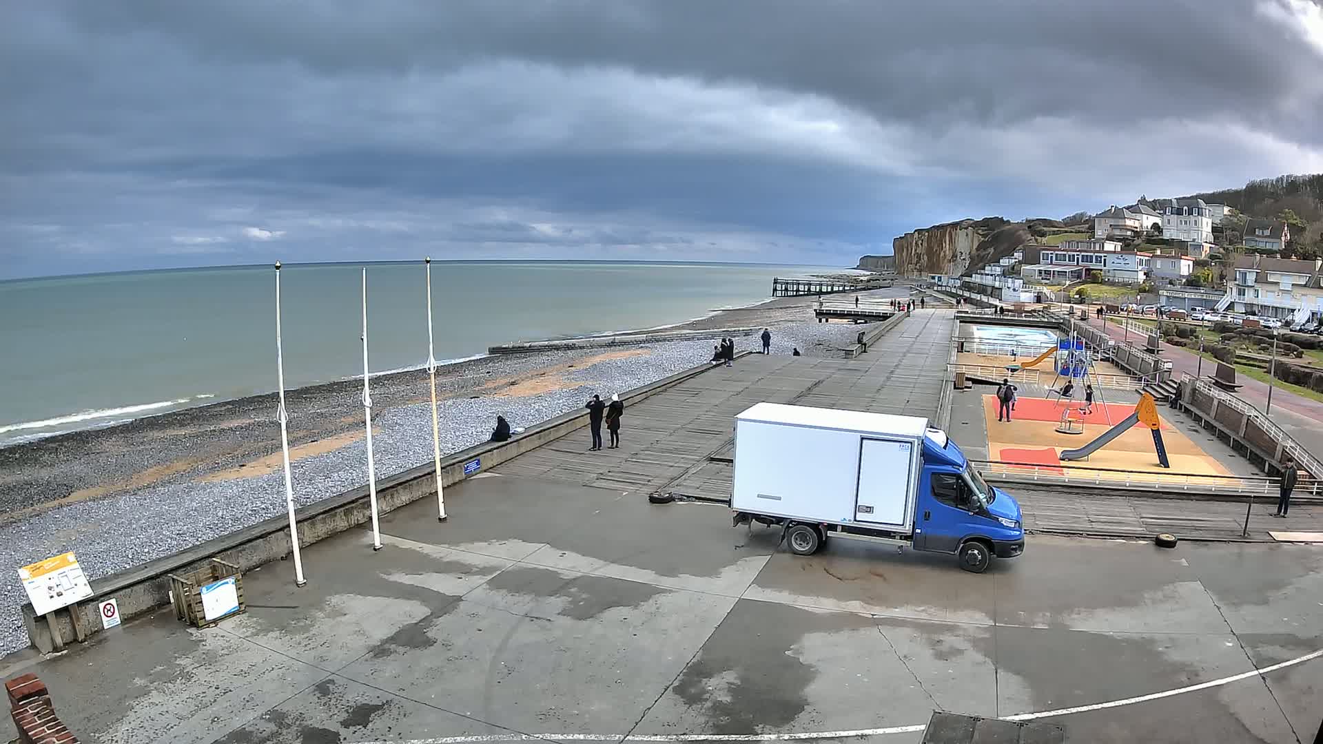 A seaside promenade with a pebble beach, a playground, a small van, and several people under a cloudy sky.