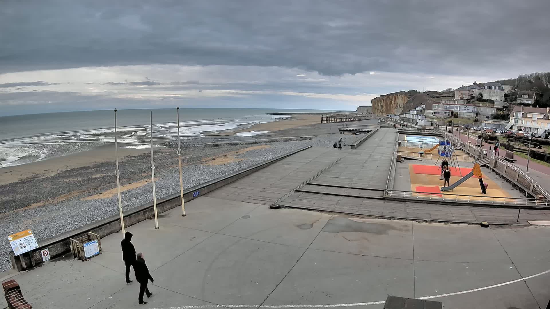 A seaside town under a cloudy sky features a paved beachfront area with a playground, a small beach with gentle waves, and a background of white buildings and cliffs.